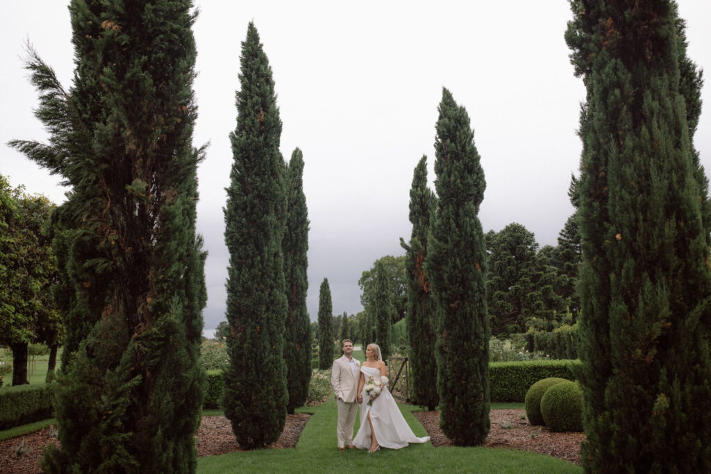 A couple stands together in a lush garden pathway flanked by tall, narrow cypress trees. The bride is wearing an off-the-shoulder white wedding dress, holding a bouquet of flowers, while the groom is dressed in a beige suit. The background features well-manicured greenery and a cloudy sky, creating a serene and elegant atmosphere.