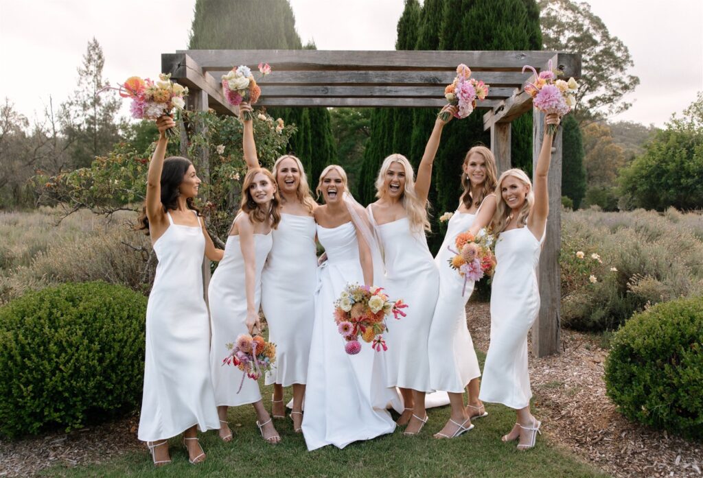 How to Enjoy Your Wedding Day Without the Worry! A group of bridesmaids, dressed in matching white mid-length dresses, happily hold bouquets up in the air while standing in a garden.